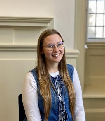 A girl with long blonde hair and glasses sits in a chair and smiles at the camera.