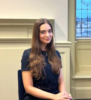 Phoebe Glynn has long brown hair and sits in a chair smiling at the camera.