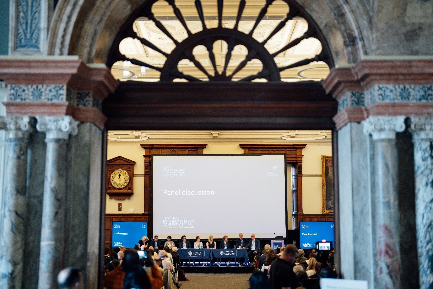 Image shows the main doors into a large room at the Examination Schools focusing on a projector screen displaying the title of the next presentation session